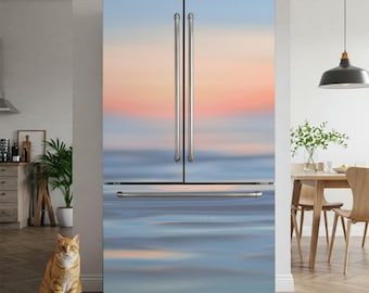 May include: A fridge with a custom wrap depicting a blurred ocean and sunset in blue, pink, and orange hues. Stainless steel handles and trim add a modern touch. An orange tabby cat sits on the wooden floor in front of the appliance.