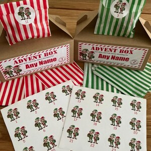 May include: Advent box set with red and white striped bags, green and white striped bags, and numbered stickers featuring elf illustrations. The boxes are brown with text that reads "Advent Box Any Name".