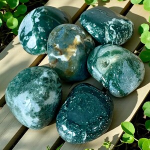 May include: A collection of five polished moss agate stones featuring deep forest green inclusions, translucent white quartz areas, and earthy brown mineral deposits. Each smooth, rounded stone displays unique natural patterns and glossy surfaces.