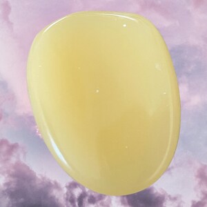 K&ouml;nnte beinhalten: Ein glatter, ovaler polierter Stein in blassgelber Farbe. Der Stein hat eine durchscheinende Optik und ist vor einem violetten und wei&szlig;en, bew&ouml;lkten Hintergrund platziert.