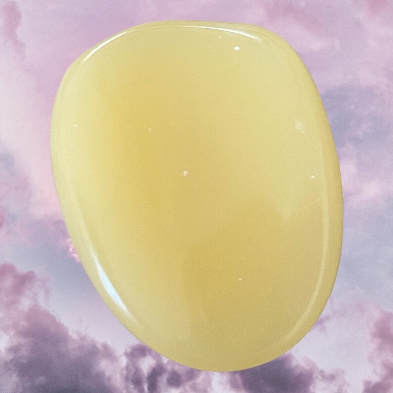 K&ouml;nnte beinhalten: Ein glatter, ovaler polierter Stein in blassgelber Farbe. Der Stein hat eine durchscheinende Optik und ist vor einem violetten und wei&szlig;en, bew&ouml;lkten Hintergrund platziert.