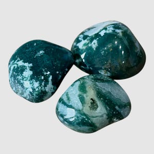 May include: Three polished moss agate stones with smooth, rounded surfaces. The stones feature deep forest green inclusions and translucent white patterns, with one stone displaying a small, textured crystalline cavity in its center.