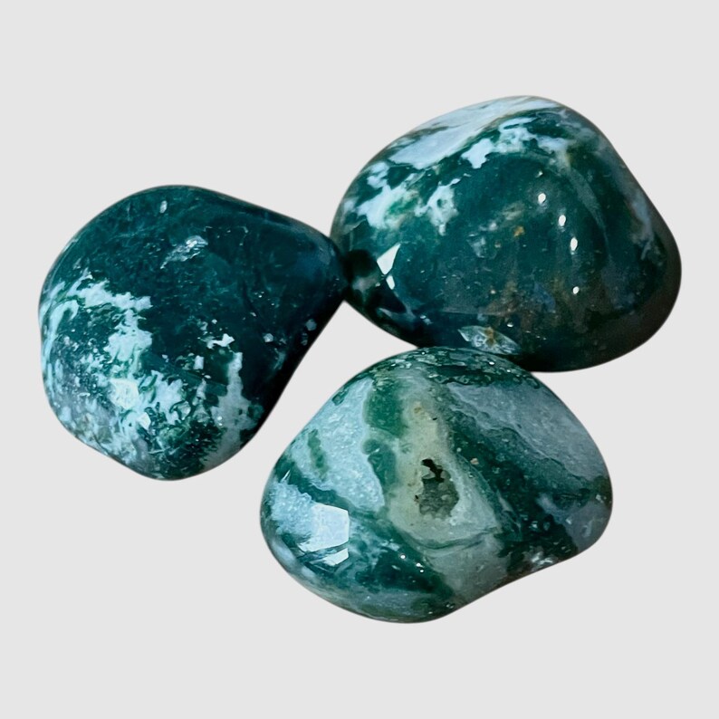 May include: Three polished moss agate stones with smooth, rounded surfaces. The stones feature deep forest green inclusions and translucent white patterns, with one stone displaying a small, textured crystalline cavity in its center.