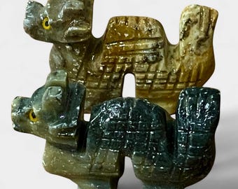 May include: Two carved stone camel figurines. The top camel is tan and brown, while the bottom camel is dark green. Both have yellow eyes and are detailed with carved patterns.