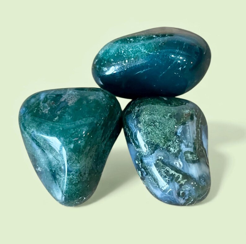 May include: Three polished moss agate stones with smooth, rounded surfaces. The stones feature deep forest green hues, translucent white inclusions, and intricate moss-like patterns throughout their crystalline structures, reflecting light.