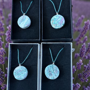 May include: Four circular pendants featuring mottled teal, green, and magenta stone patterns, each suspended from a dark teal cord. The pendants are individually displayed in black velvet-lined boxes, showcasing natural mineral textures and silver bails.
