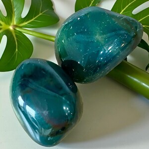 May include: Two polished, smooth, rounded stones with a deep teal and blue-green color palette. The stones feature translucent areas and internal veining, displaying a glossy, reflective surface that catches the light, highlighting their organic shape.
