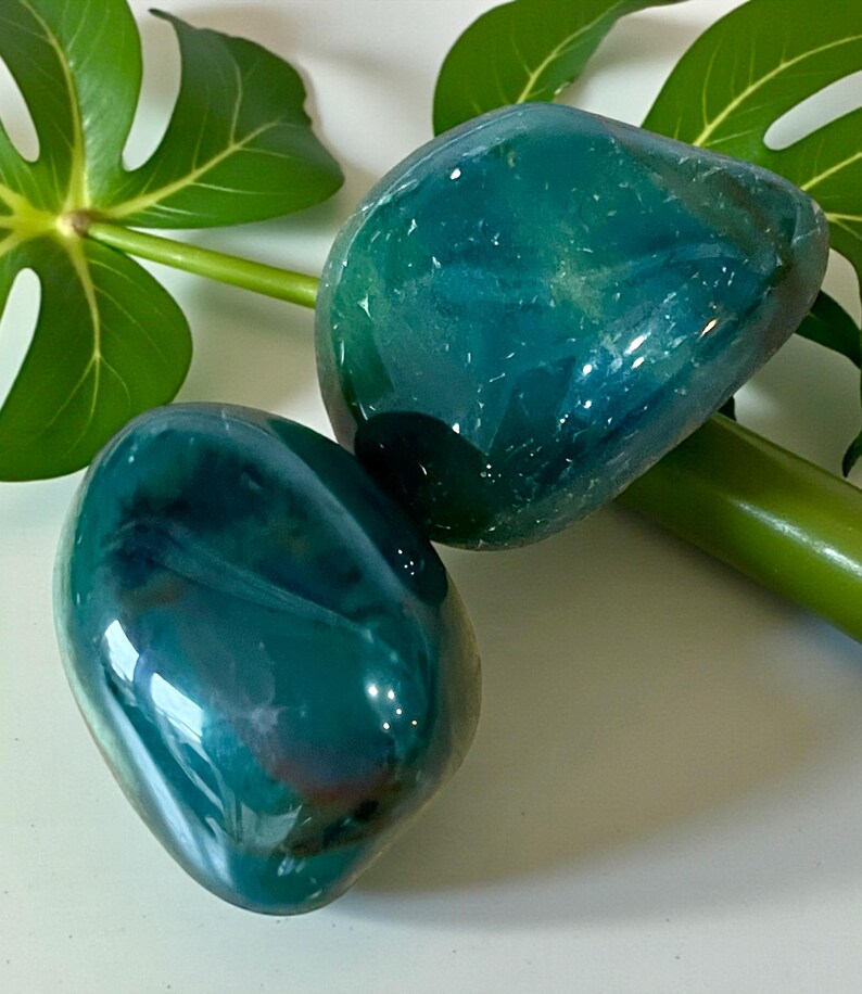 May include: Two polished, smooth, rounded stones with a deep teal and blue-green color palette. The stones feature translucent areas and internal veining, displaying a glossy, reflective surface that catches the light, highlighting their organic shape.