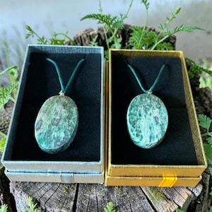 May include: Two oval gemstone pendants on green cords, displayed in open gift boxes. The pendants feature a mottled green and red-brown pattern. One box is silver, the other gold, both with black interiors. The boxes rest on a weathered wooden surface.