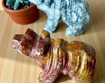 May include: Two carved stone bear figurines. One is a mix of red, brown, and yellow hues, while the other is gray. A small potted succulent plant is in the background. The bears are detailed and appear to be handcrafted.