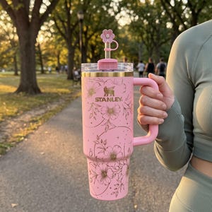 Puede incluir: Un vaso Stanley de color rosa claro con asa, que presenta un patrón de líneas florales doradas, una tapa transparente y una pajita de metal rematada con una cubierta de silicona en forma de flor rosa. La marca Stanley aparece en dorado.
