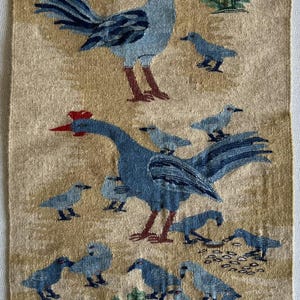 Op de afbeelding: Een beige kleed met een geweven ontwerp van een haan en een gans met hun kuikens. De haan is blauw en rood, en de gans is blauw en rood met een lange nek. De kuikens zijn allemaal blauw.