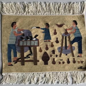 May include: A woven tapestry depicting three figures working with pottery. The figures are dressed in blue and brown clothing. The tapestry is beige with a white fringe border. The tapestry has the text "oita" in the bottom right corner.