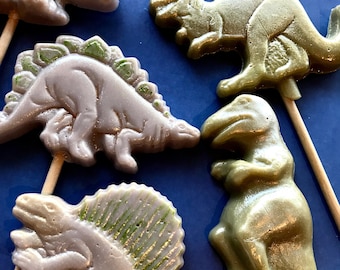 May include: Four dinosaur-shaped lollipops on sticks. Two are green with a shimmery finish and two are purple with a shimmery finish. The dinosaurs are all different types, including a stegosaurus, a tyrannosaurus rex, and a triceratops.