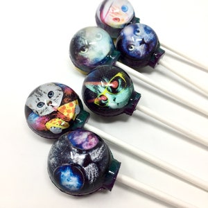 May include: A set of six colorful lollipops with different cat-themed designs. The lollipops have white sticks and are arranged in a row on a white surface.