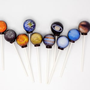 May include: A set of nine colorful lollipops, each decorated with a different planet from the solar system. The lollipops are arranged in a semi-circle on a white background.