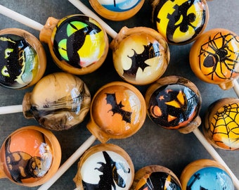 May include: A collection of Halloween-themed lollipops with various designs, including bats, spiders, pumpkins, and witches. The lollipops are on wooden sticks and have a clear, glossy finish.