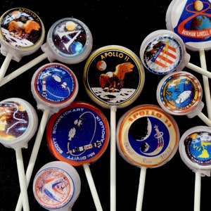 May include: A collection of 11 round, colorful lollipops with images of the Apollo missions. Each lollipop features a different mission patch with the mission number and crew names. The lollipops are on white sticks.
