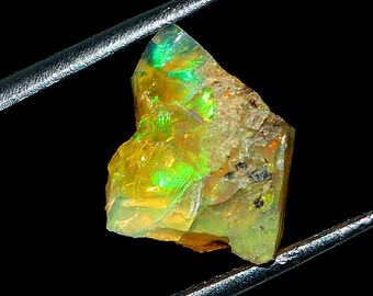 May include: A rough, green and yellow opal gemstone with iridescent flashes. The opal is held in tweezers against a black background.