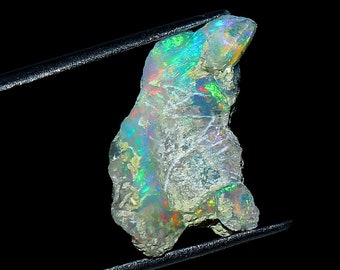 May include: A rough, white opal with iridescent green, blue, and red flashes. The opal is held by tweezers against a black background.