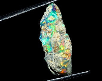 May include: A rough, iridescent opal gemstone with a green, blue, and orange play of color. The opal is held by a pair of tweezers against a black background.