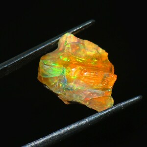 Peut inclure: Une opale brute non taillée, tenue par une pince. L'opale présente un jeu de couleurs vibrant, avec des éclats d'orange, de vert et de bleu. La pierre a une surface texturée et une forme irrégulière, sur fond sombre.