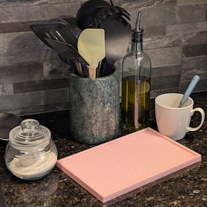 以下が含まれることがあります： テクスチャ加工された表面を持つピンクのシリコン製ベーキングマット、蓋付きのガラス瓶、白いセラミック製のマグカップ、コルク栓付きのガラス瓶、黒いキッチン用品が入った石製のツールホルダー