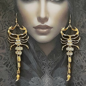 Può includere: Orecchini a forma di scorpione color oro con dettagli in cristallo trasparente. Gli orecchini sono esposti davanti a un ritratto di donna con capelli scuri e un copricapo decorativo. Chiusura a gancio.