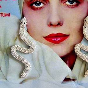 Può includere: Una copertina vintage della rivista Vogue Italia. L'immagine mostra una donna con rossetto rosso e orecchini di strass a forma di serpente. Il titolo della rivista è in rosso, con la parola "Italia" sopra.