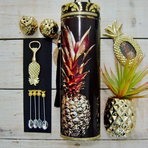 May include: A selection of gold pineapple-themed items on a white wooden surface. Features a tall, cylindrical container with a pineapple design, a pineapple bottle opener, salt and pepper shakers, and small spoons. A pineapple picture frame and a small gold pineapple vase are also present.