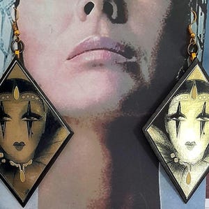 Könnte beinhalten: Ein Paar rautenförmige Ohrringe mit einem stilisierten Gesicht und goldenen Akzenten. Das Gesicht hat dunkle Augenmarkierungen und eine goldene Krone. Die Ohrringe hängen an goldfarbenen Haken. Der Hintergrund ist ein verschwommenes Bild eines Gesichts.