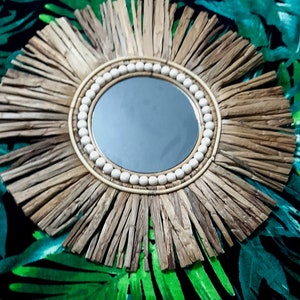 May include: Round decorative mirror with a natural, sunburst-style frame. The frame is made of light brown, textured material, radiating outwards from a central circular mirror. A ring of small, white beads surrounds the mirror's edge.