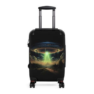 May include: Black rolling suitcase featuring a vibrant illustration of a UFO hovering over a forest landscape. The artwork includes a bright green beam of light, a road, and a person walking. The suitcase has a retractable handle and four wheels.