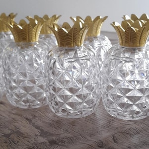 Puede incluir: Una colección de recipientes transparentes de vidrio en forma de piña con tapas doradas. Los recipientes tienen un patrón de corte de diamante. Las tapas tienen forma de coronas de piña. Los objetos están dispuestos sobre una superficie de madera.