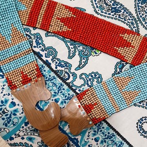 May include: A beaded belt with a wooden buckle. The belt features a pattern of red, tan, and light blue beads. The wooden buckle is a light brown colour. The belt is displayed on a paisley patterned fabric.