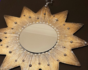 May include: A star-shaped mirror with a gold and silver finish. The mirror is surrounded by a border of white pearls and silver beads. A small silver star and a silver wire are attached to the top for hanging.