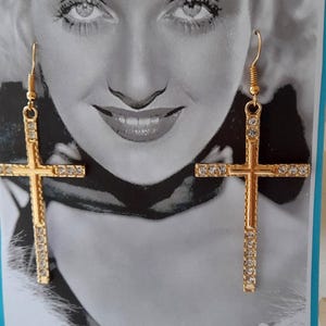 May include: Gold-coloured cross earrings with clear crystal detailing. These earrings have a traditional cross shape and a hook fastening. The crystals are set along the arms of the cross, providing a sparkling effect. The earrings are set against a monochrome background.