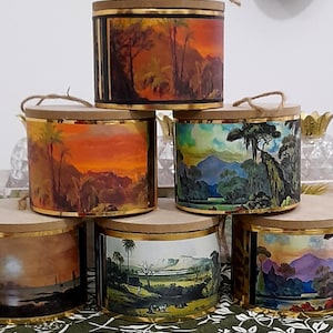 May include: A selection of cylindrical storage tins with lids. Each tin is decorated with a landscape painting in warm tones, gold trim, and a jute rope handle. The landscapes depict trees, mountains, and sunsets.