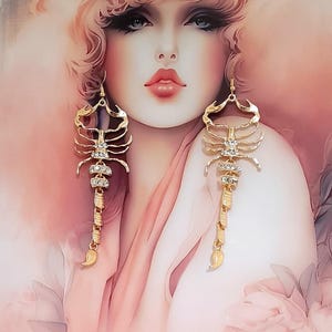 May include: Gold-coloured scorpion earrings with clear crystal accents. The earrings feature a detailed scorpion design with a long, dangling tail. The background is a soft pink and peach colour.