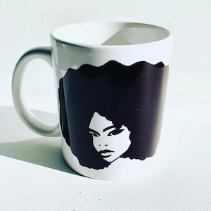May include: White ceramic mug with a black and white silhouette of a woman with an afro hairstyle.