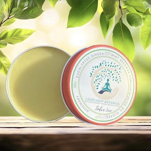 May include: A round tin of salve with a light green balm next to a tin with a red rim and a white label. The label features a tree design with a person meditating. Text includes "Eucalyptus Ginger" and "Lovelight Botanica Salve 57g."