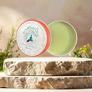 May include: A round, open tin of Lovelight Botanica salve with a light green balm, next to a tin lid with text and a tree design. The tins are on a stone platform, surrounded by greenery and flowers.