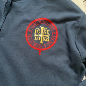 May include: Navy blue hooded sweatshirt with a full zipper. The sweatshirt features a red and gold circular emblem with religious text and a cross. The text reads "LORD JESUS CHRIST HAVE MERCY ON ME".