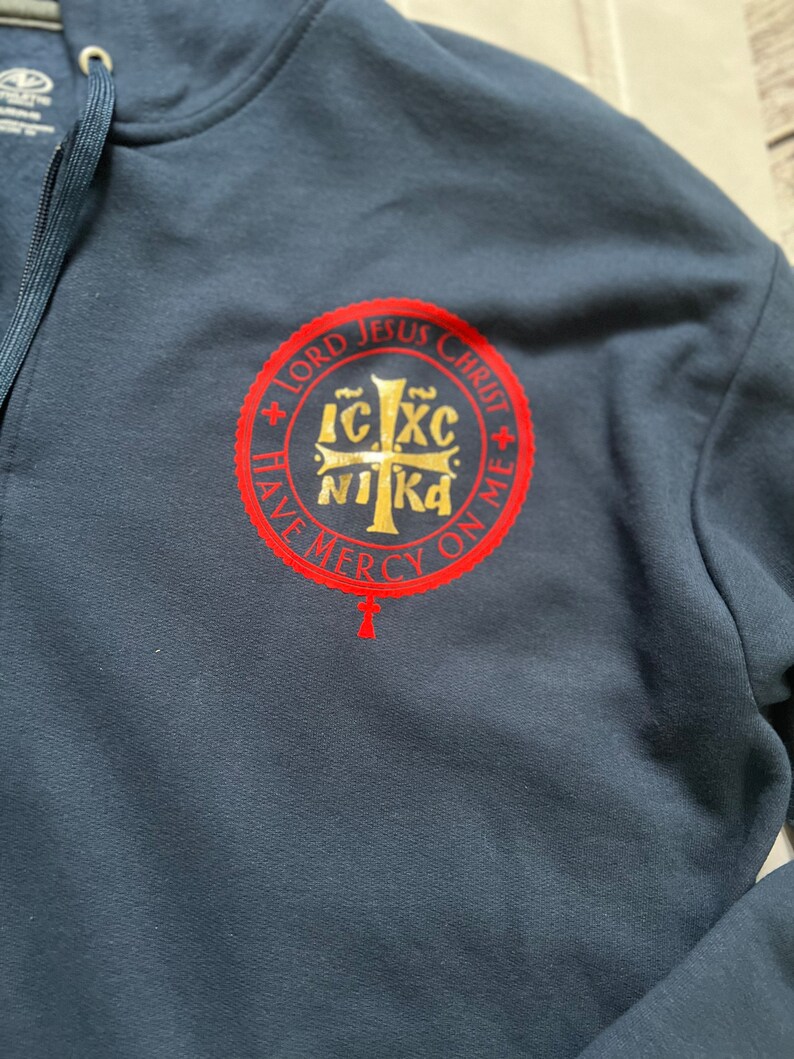 May include: Navy blue hooded sweatshirt with a full zipper. The sweatshirt features a red and gold circular emblem with religious text and a cross. The text reads "LORD JESUS CHRIST HAVE MERCY ON ME".