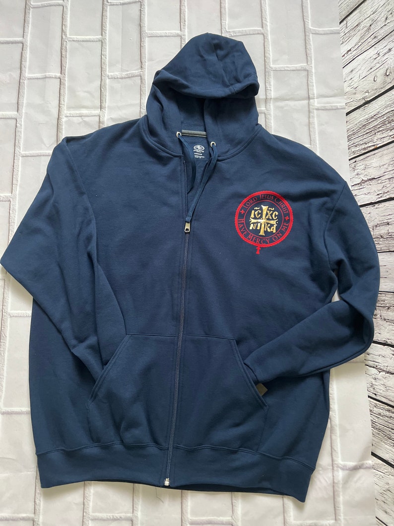May include: Navy blue zip-up hoodie with a hood and front pockets. A circular emblem with text in red and gold is on the left chest. The hoodie is made of a soft material.