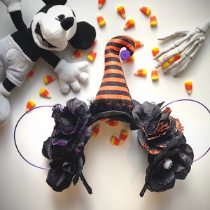 May include: Black and orange witch hat headband with black flowers and a purple headband. The headband has a single black eye on one of the flowers.