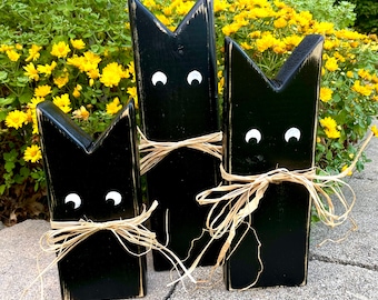 May include: Three black wooden cat figurines with white eyes and a straw bow. The cats are different sizes and are arranged in a row on a gray surface.