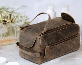 May include: A brown leather toiletry bag with two zippered compartments and a top handle. The bag has a distressed finish and a flap closure. The bag is sitting on a marble surface.