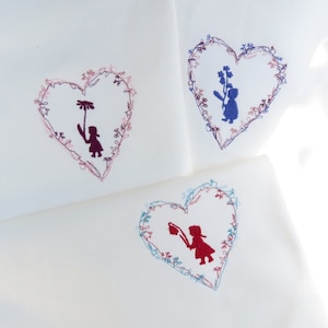 Puede incluir: Tres paneles de tela blanca con corazones bordados. Cada corazón presenta un diseño floral y una silueta de una persona sosteniendo una flor. Los corazones están en tonos de rosa, azul y rojo.