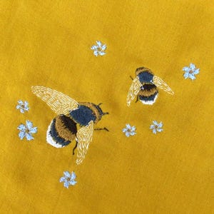 Könnte beinhalten: Senfgelber Stoff mit gestickten Bienen und kleinen blauen Blumen. Zwei Bienen sind mit goldenen, schwarzen und weißen Details bestickt. Der Stoff hat ein strukturiertes Aussehen und ist hell beleuchtet.